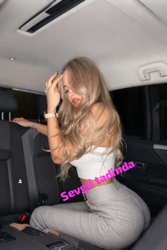 6a24291078445923547805 - Gebze Escort Blonde woman in a white top and gray pants sits in the car's back seat, touching her hair; pink caption 'Sevgili tadında' on image.