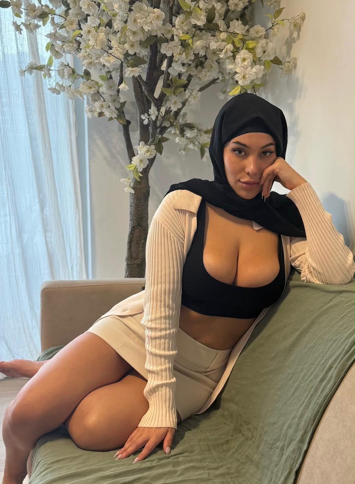 6a6fb8dbcb3d7782719107 - Gebze Escort Woman wearing a black hijab and beige cardigan sits on a green couch, resting her head on her hand with a white flower tree in the background.